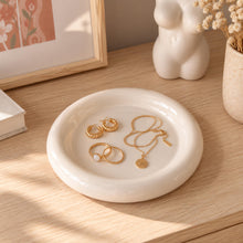 Charger l'image dans la galerie, Coupelle à bijoux blanche contenant des bagues, boucles d'oreilles et un collier dorés sur une commode en bois clair
