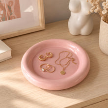 Charger l'image dans la galerie, Coupelle à bijoux rose contenant des bagues, boucles d'oreilles et un collier dorés sur une commode en bois clair
