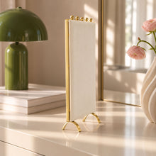 Charger l'image dans la galerie, Porte-colliers vide en velours écru et métal doré sur une commode, près d'une lampe verte et de fleurs roses
